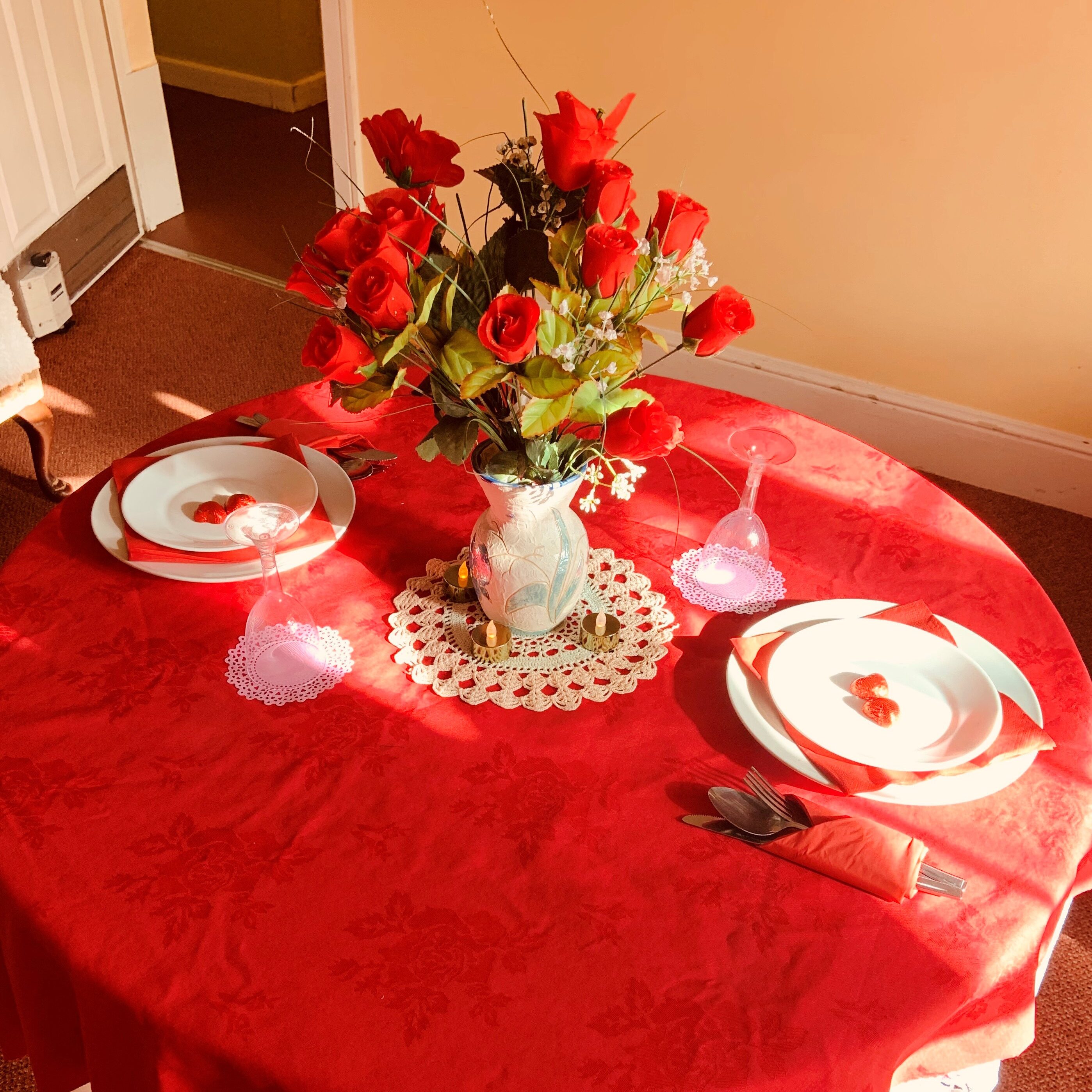 Shows a table laid for two. Red tablecloth, white dinner service with red napkins and a beautiful flower display of roses in crystal glass with white doiley.